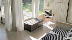Een kamer met een houten vloer, een gestreept bankje, een rieten stoel, een klein tafeltje en een raam met gordijnen.