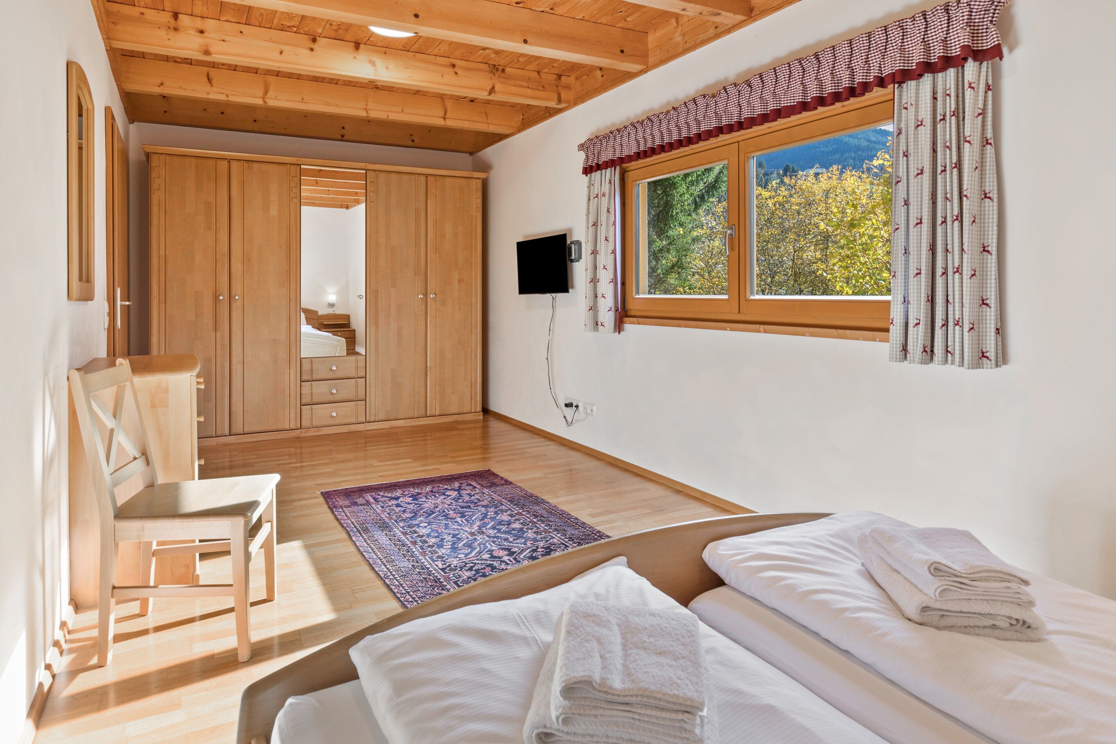 Une chambre avec un plafond en bois, un lit recouvert de draps blancs, une fenêtre avec des rideaux, une armoire en bois et une chaise.