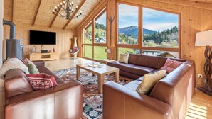 Ein Holzinnenraum mit einer Couch, einem Couchtisch und großen Fenstern mit Blick auf eine gebirgige Landschaft.