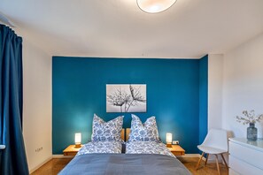 A bedroom with a bed, two bedside tables with lamps, a white chair, a desk with a vase, and a wall art piece.