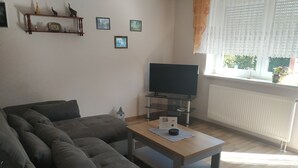 A living room with a sofa, a TV on a stand, a small table, and a window with curtains. - "Kijas" holiday flat with private terrace and WLAN (Wismar)