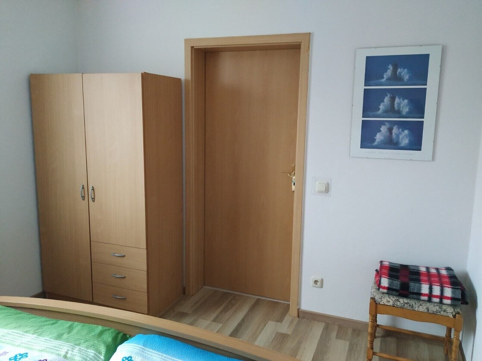 A bedroom with a wardrobe, a bed, a small stool, and a wall-mounted picture.