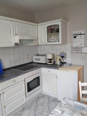 Uma cozinha compacta com armários brancos, micro-ondas, cafeteira e uma mesa pequena com toalha de mesa florida.