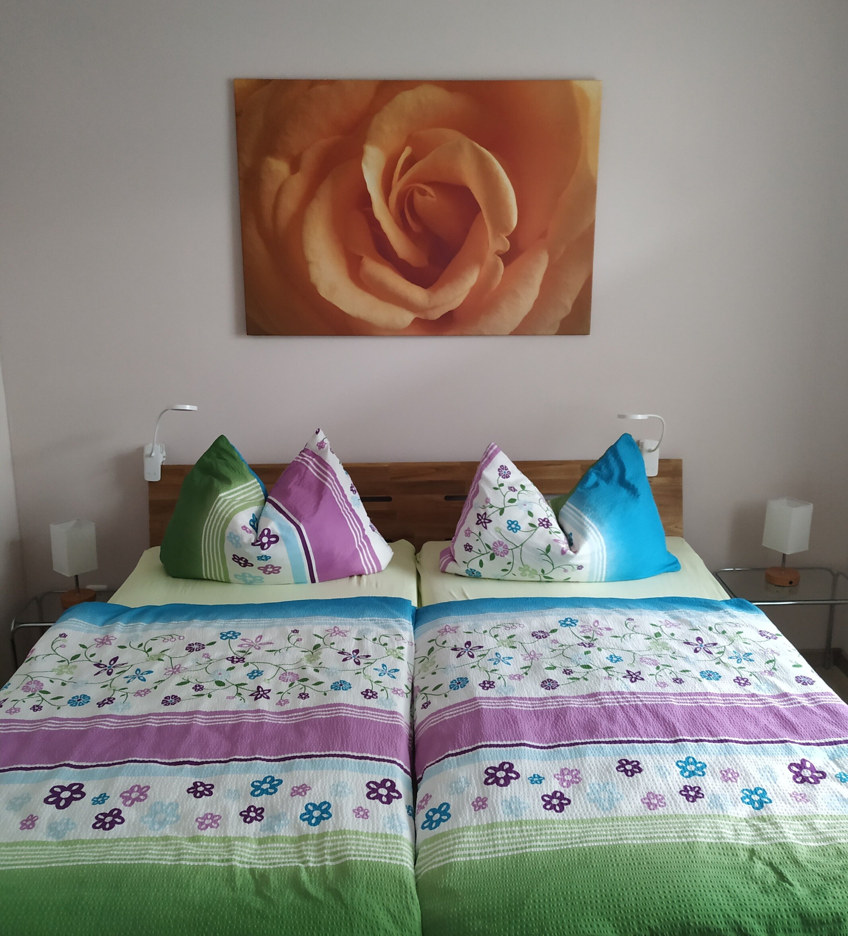 A double bed with floral-patterned bedding and two pillows, a wall-mounted painting of a rose, and bedside lamps.