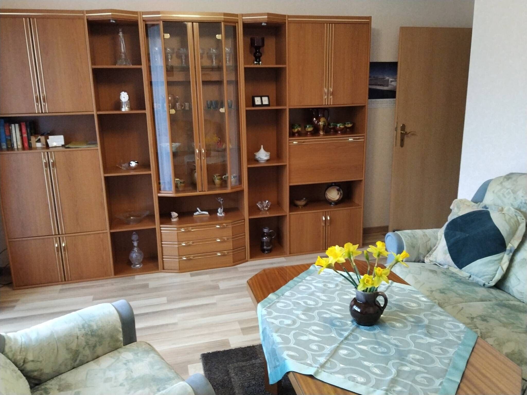 A living room with a wooden cabinet, a sofa, a coffee table with a vase of flowers, and a patterned tablecloth.