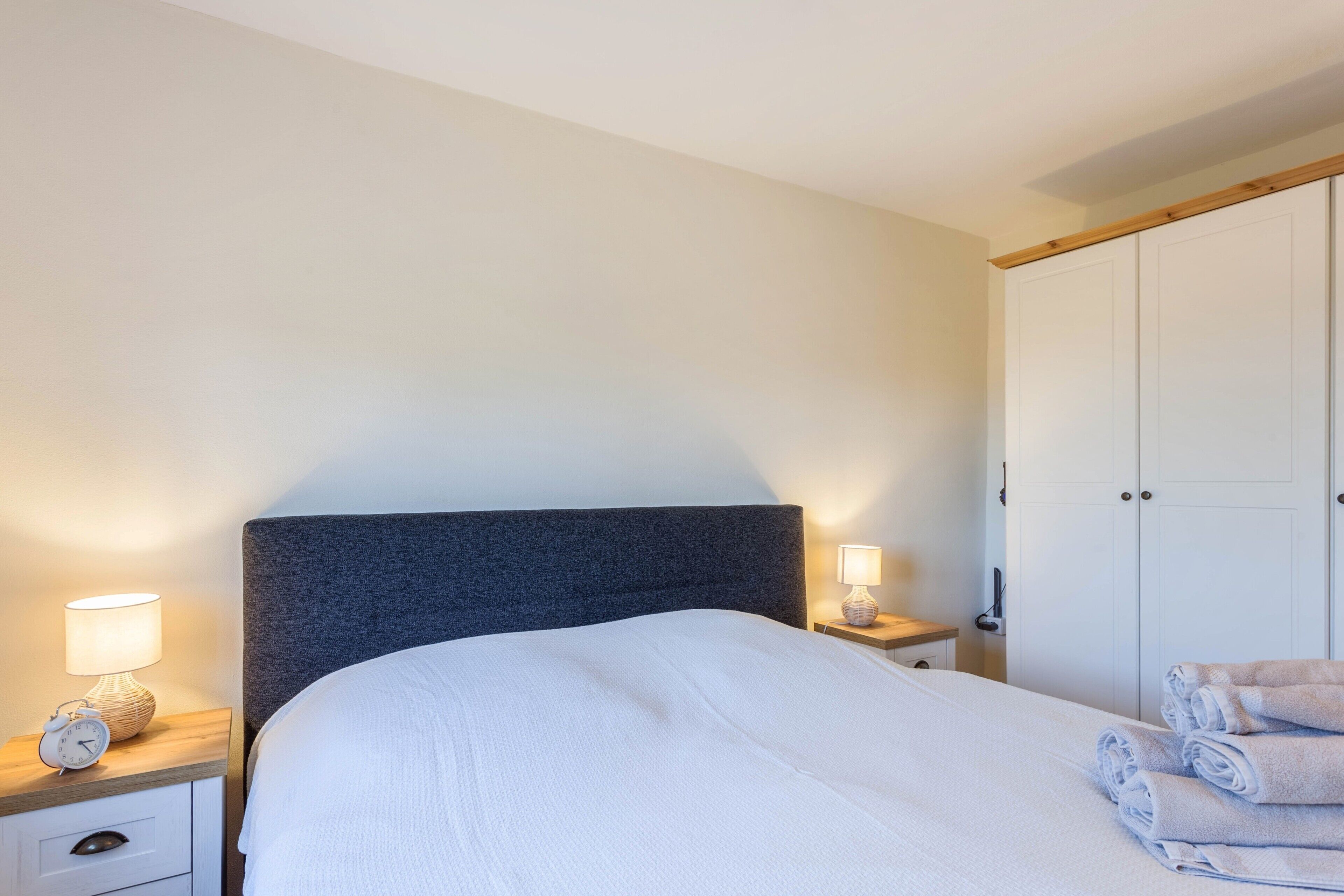 A neatly made bed with a dark headboard, a bedside table with a lamp, and a wardrobe with a wooden top.