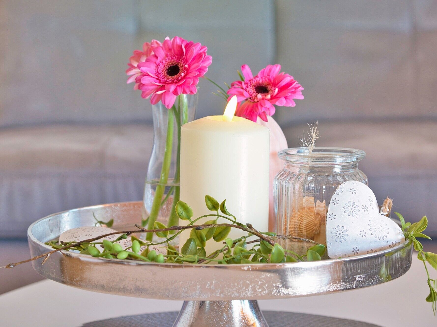 A candle, two pink flowers, a jar with seashells, and a heart-shaped object on a tray.
