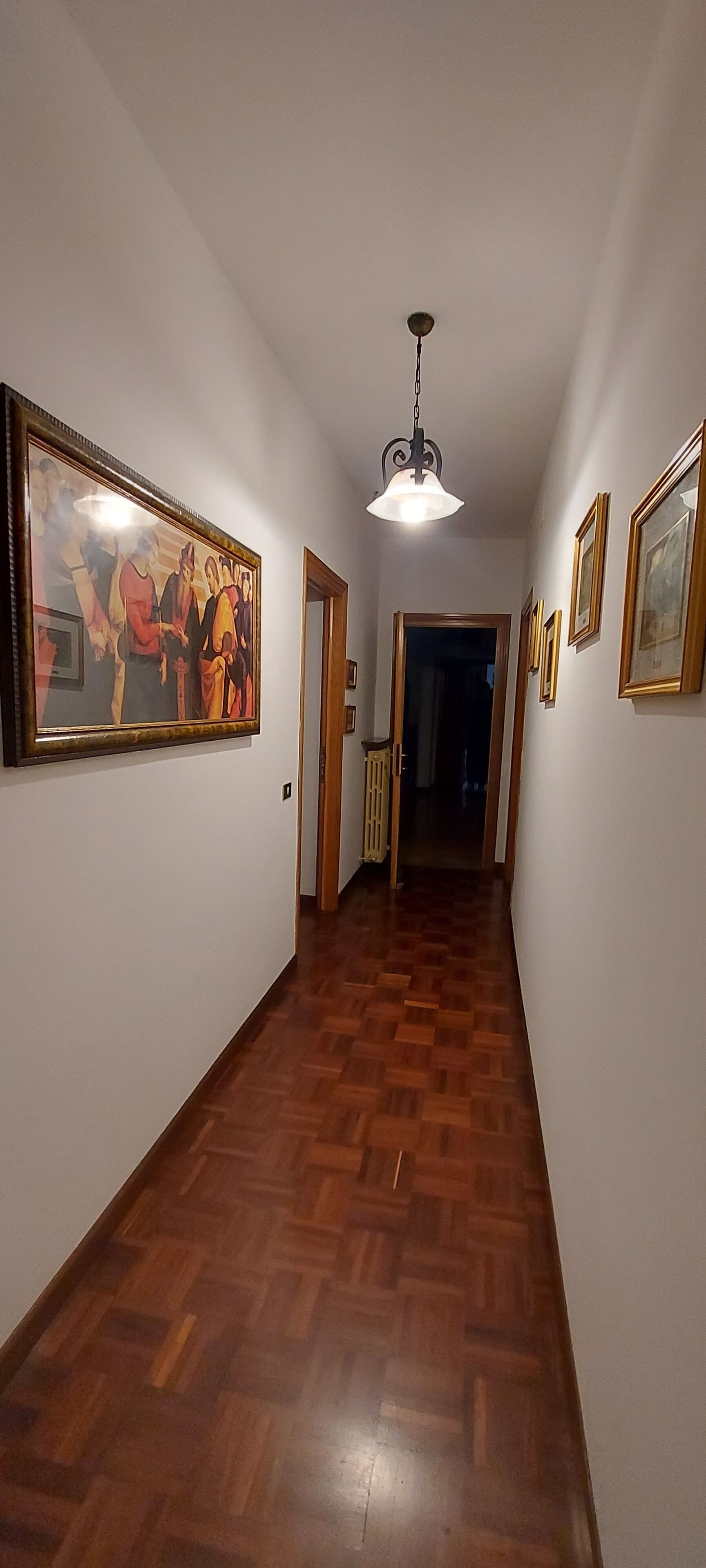 A hallway with wooden flooring, framed artwork on the walls, and a hanging light fixture.