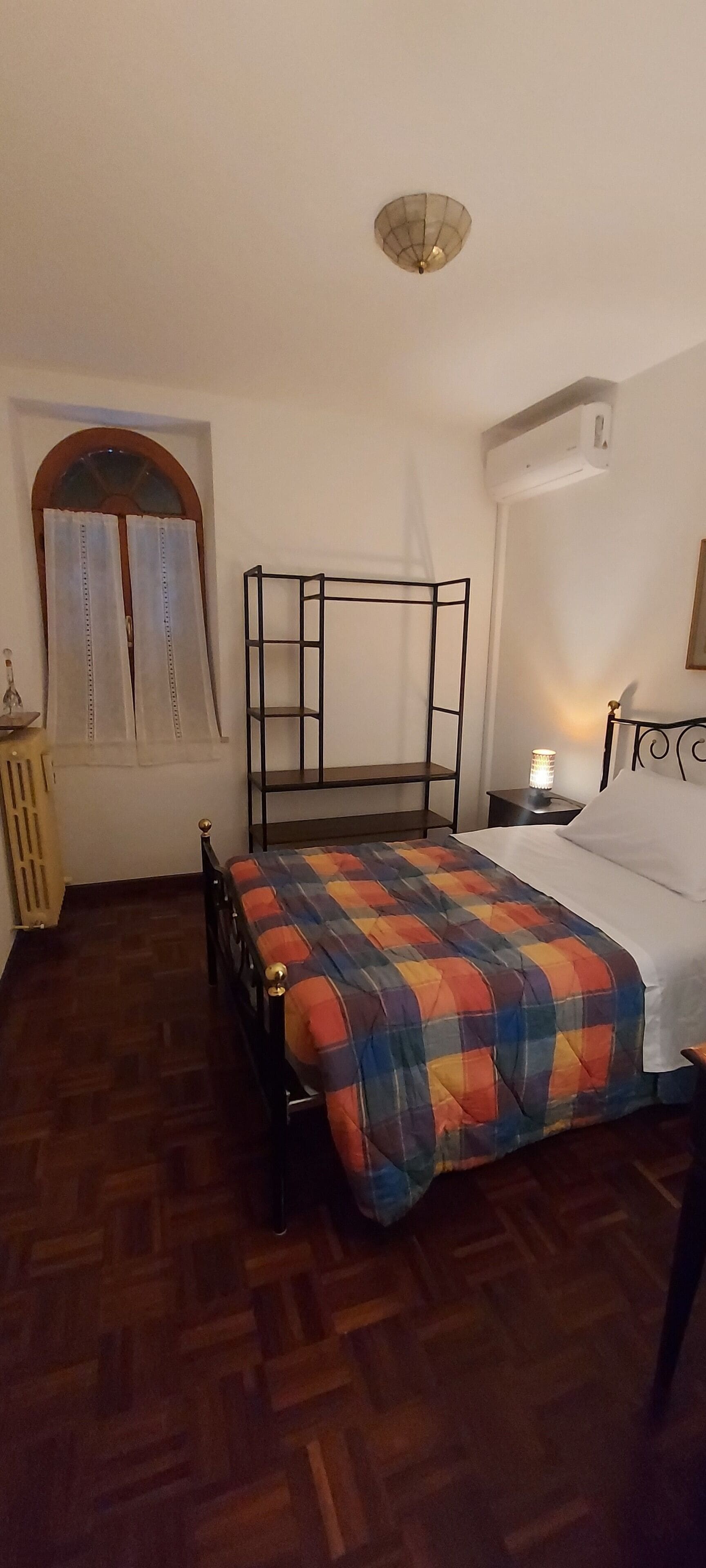 A bedroom with a bed, a bedside table with a lamp, a window with curtains, and a radiator.