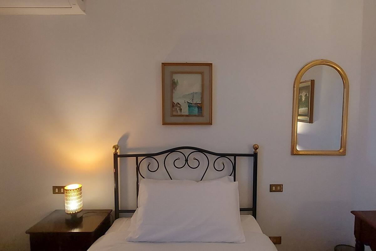 Un lit bien fait, avec une tête de lit, une lampe de chevet, un tableau encadré au mur et un miroir.