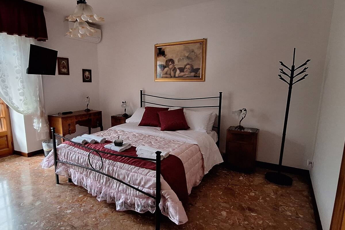 Une chambre  à coucher avec un lit, une table de chevet, une lampe, un tableau et une fenêtre avec des rideaux.