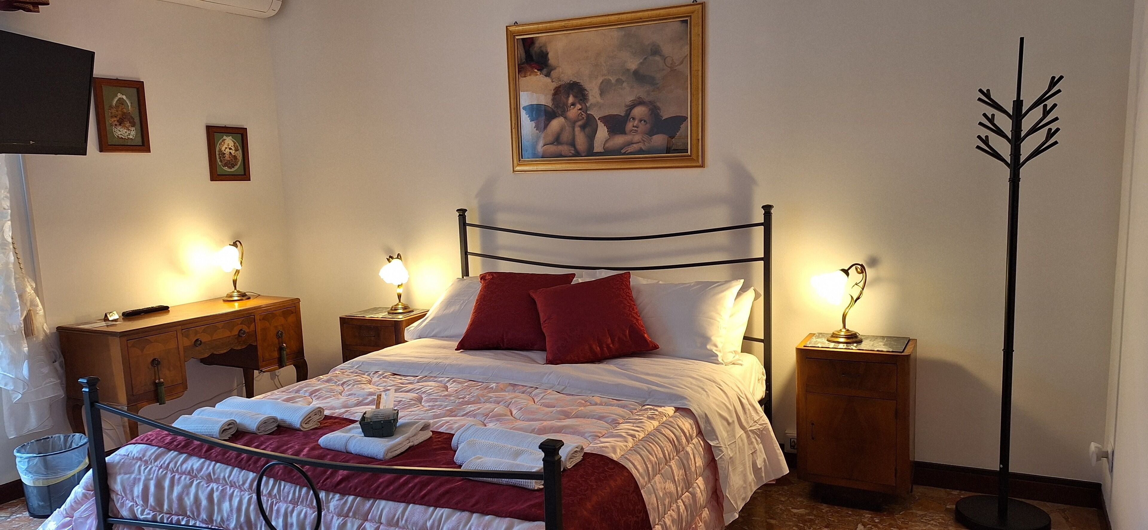 A neatly made bed with red and white linens, two bedside tables with lamps, a painting on the wall, and a floor lamp.