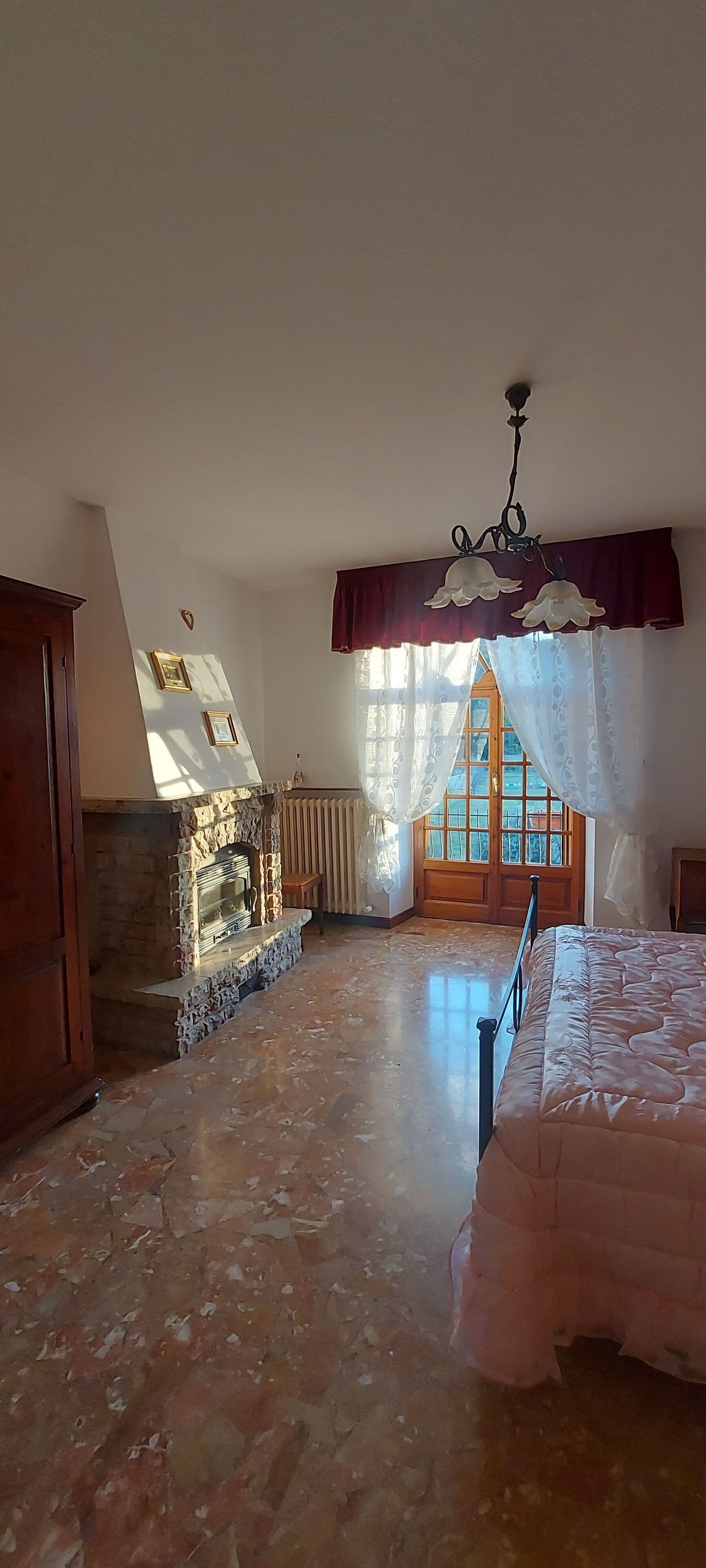 A bedroom with a stone fireplace, a bed, a wooden wardrobe, and a window with curtains.