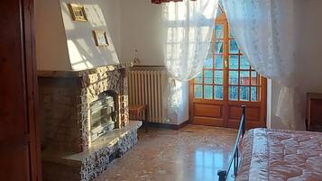 A bedroom with a stone fireplace, a bed, a wooden wardrobe, and a window with curtains.