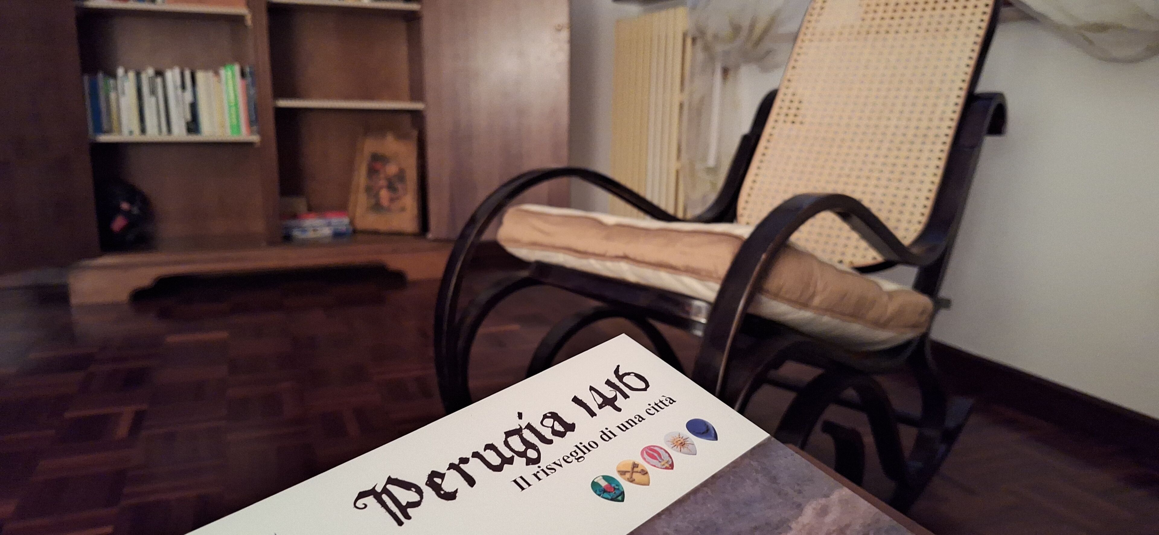 Una stanza con pavimento in legno, una libreria, una sedia in vimini e un tavolo con un cartello.