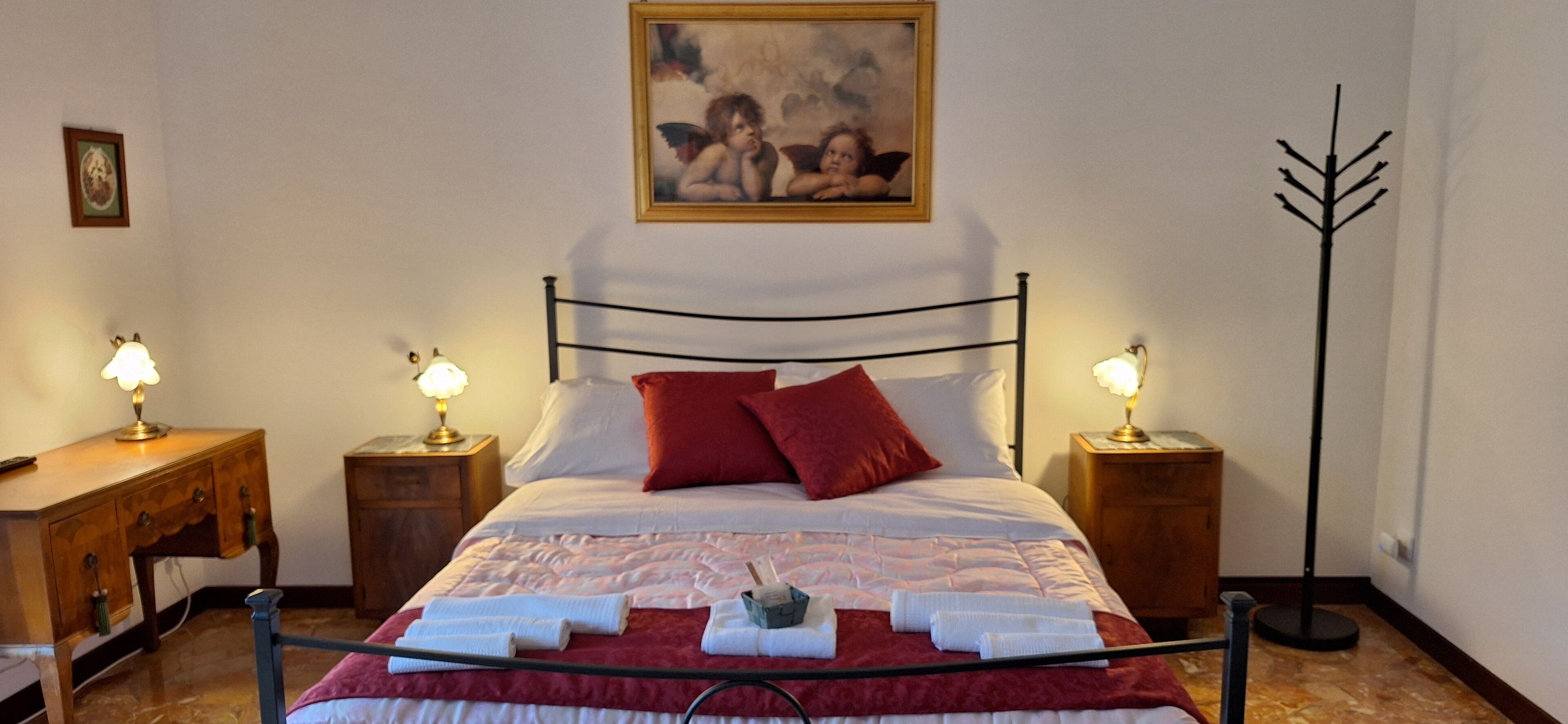 Un lit bien fait, avec du linge de lit rouge et blanc, deux tables de chevet avec des lampes, un bureau en bois et un tableau accroché au mur.