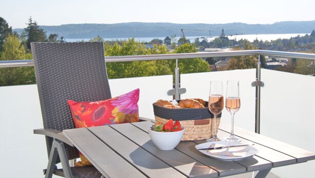 Een tafel met een rieten stoel, een mand met brood, een schaal aardbeien en twee glazen wijn op een balkon met uitzicht op een meer.
