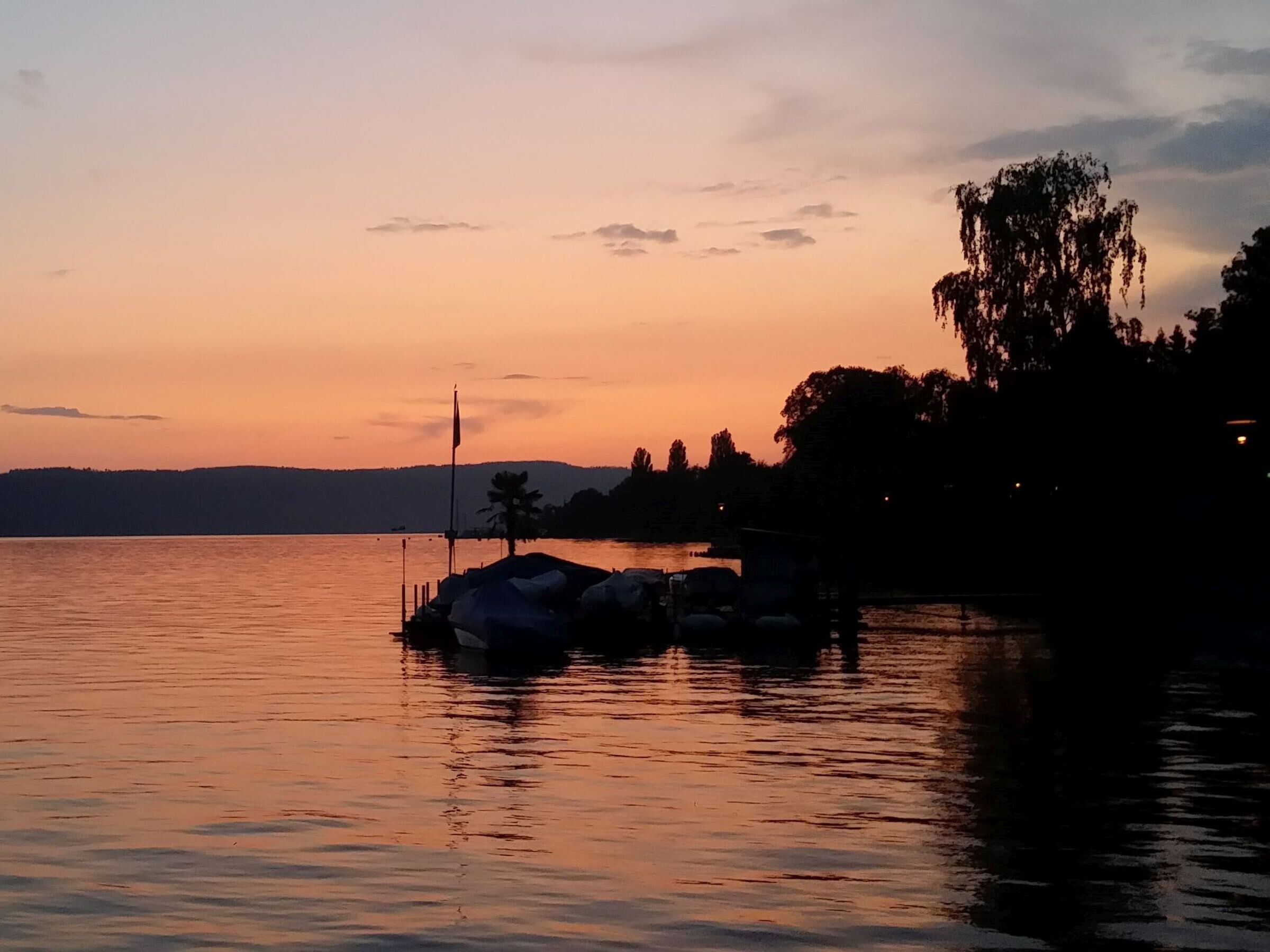 Ein ruhiger See bei Sonnenuntergang mit einem Steg und Bäumen im Hintergrund.