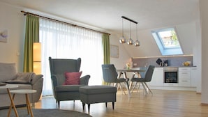 Een moderne woonkamer met een grijze bank, een bijpassende fauteuil, een eettafel met stoelen, een keukenhoek met raam en dakraam.