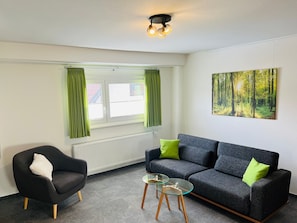 A living room with a grey sofa, a black armchair, a glass coffee table, and a window with green curtains.