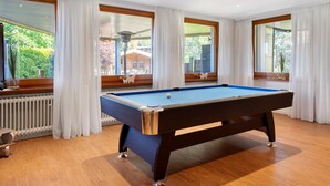 Una mesa de billar en una habitación con amplios ventanales, suelo de madera y un radiador.