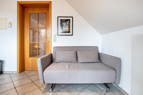Una habitación con un sofá gris, una puerta de madera con paneles de vidrio, un cuadro en la pared y suelo de baldosas.