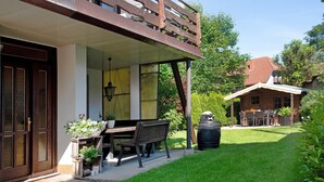 Une maison avec une véranda, un banc en bois, une table à manger et un grand tonneau.