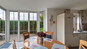 A dining area with a table set for two, a vase with flowers, and a kitchen with a refrigerator and sink. - Holiday Home 'Ferienhaus Beckerwitz' with Private Terrace, Garden & Wi-Fi (Beckerwitz)