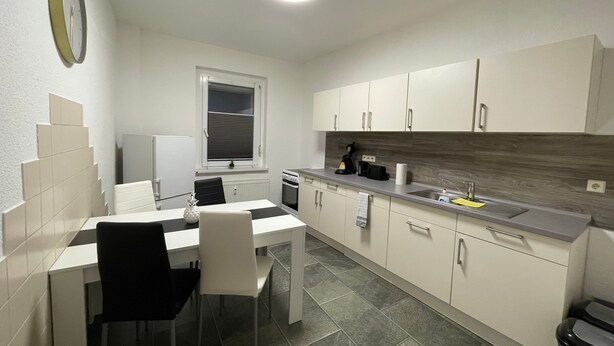 A compact kitchen with white cabinets, a dining table with chairs, a built-in oven, and a window with blinds. - Apartment 'Das Grüne Haus' with Shared Terrace and Shared Garden (Finsterwalde)