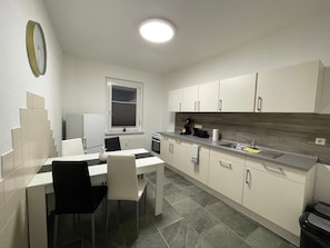 Een kleine keuken met witte kastjes, een eettafel met stoelen, een inbouwoven en een raam met zonwering.
