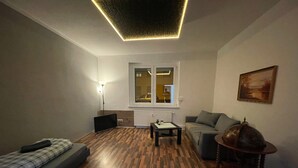 A living room with a sofa, a coffee table, a lamp, a TV, and a window with a view of the outside. - Apartment 'Das Grüne Haus' with Shared Terrace and Shared Garden (Finsterwalde)