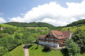 A large, multi-story building with a sloped roof, surrounded by lush green hills and trees.