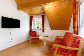 Et værelse med et fjernsyn ophængt på væggen, trælofter, et vindue med gardiner, en rød sofa med et mønster, en træstol og et lille rundt bord.