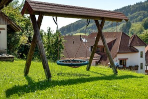 Legehus i træ med gynge, omgivet af frodig græs og et landligt landskab med huse.