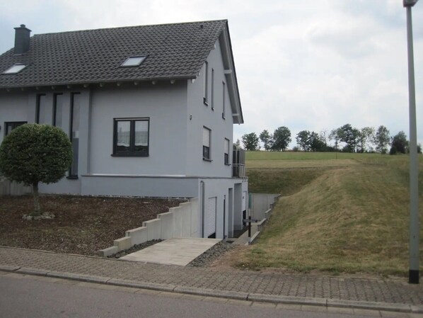 A modern two-story house with a grey facade, a small front yard with a trimmed bush, and a sloped roof.
