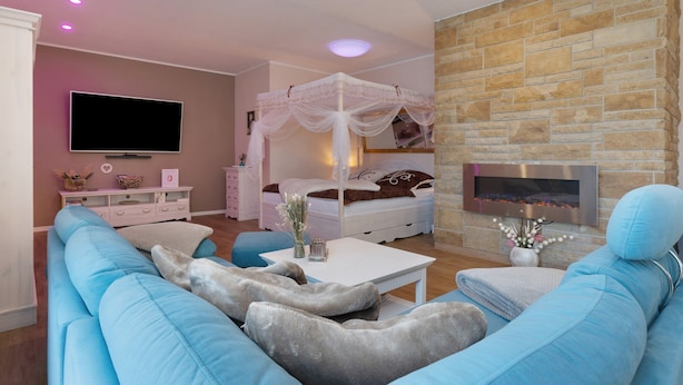 A modern living room with a sofa, a television, a white bedside table, a white canopy bed, and a stone fireplace.