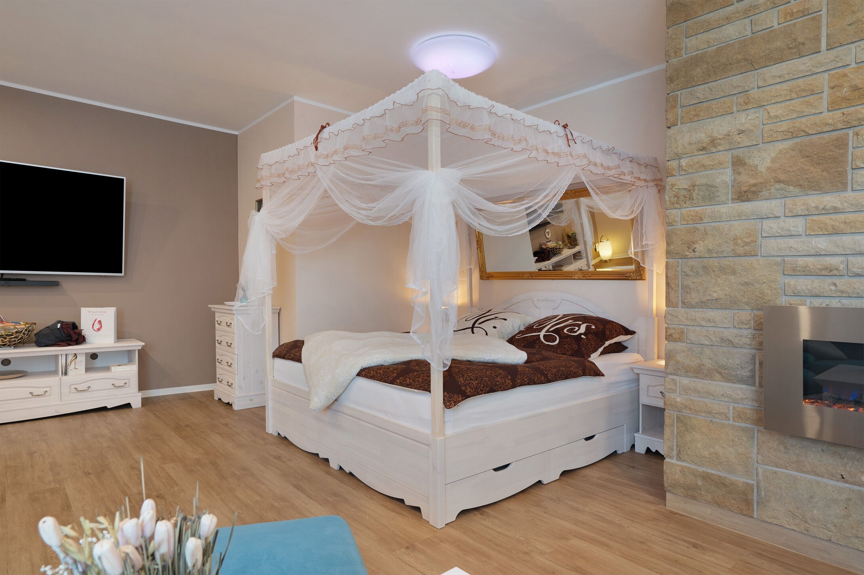 A bedroom with a four-poster bed, a flat-screen TV, a stone wall, and a fireplace.
