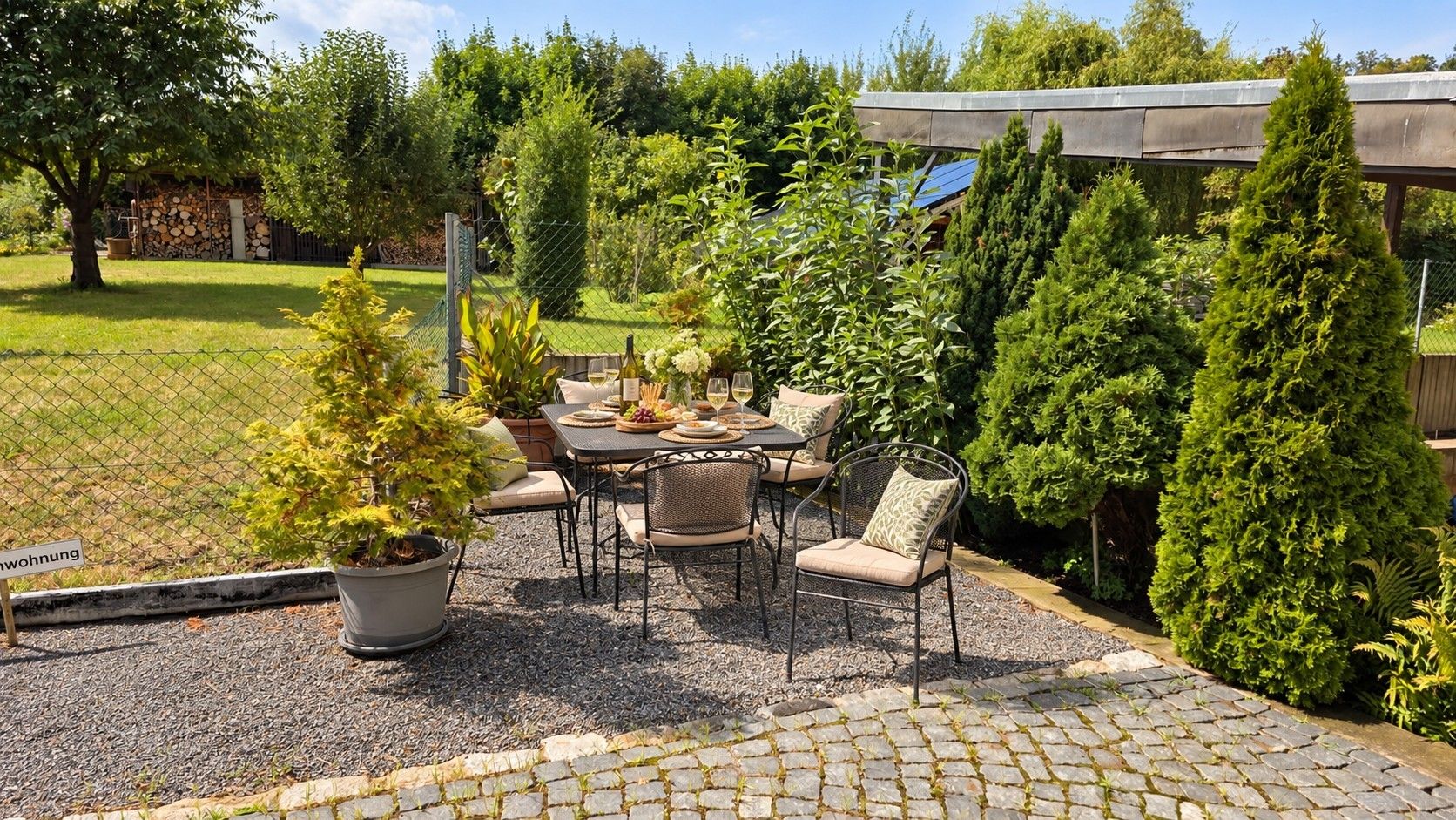 Uma área de pátio ao ar livre com mesa e cadeiras, cercada por diversos vasos de plantas e um jardim murado.
