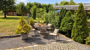 Ein Terrassenbereich im Freien mit Tisch und Stühlen, umgeben von verschiedenen Kübelpflanzen und einem eingezäunten Garten.
