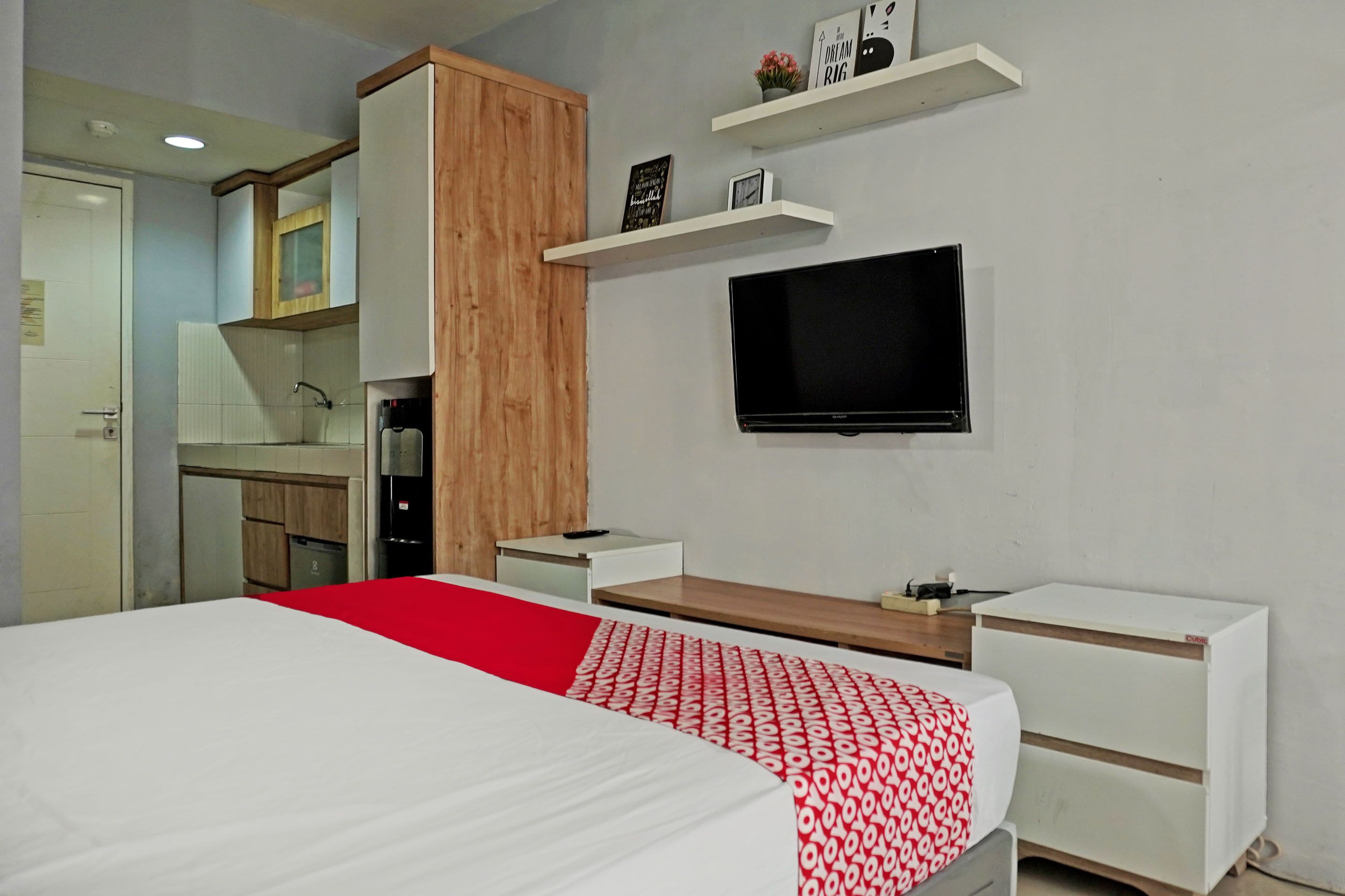 Een slaapkamer met een bed, een televisie, een houten kast, een kitchenette en een sprei met een rood-wit patroon.
