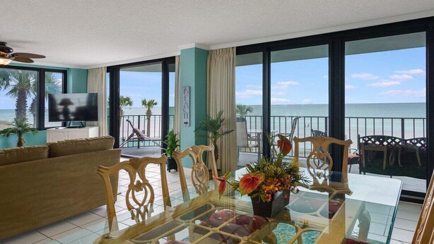 A spacious living room with a large glass table, comfortable seating, a flat-screen TV, and a balcony with a view of the sea. - Watercrest 203 - FREE Beach Chairs! (Panama City Beach)