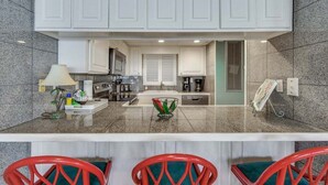 A compact kitchen with white cabinets, a central island with chairs, and a dining area with red chairs. - Watercrest 203 - FREE Beach Chairs! (Panama City Beach)