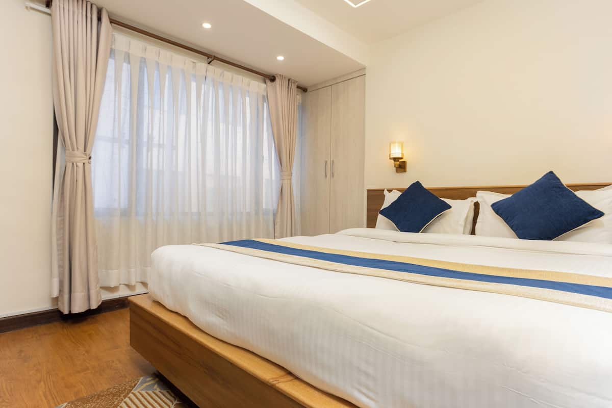 Quarto de hotel com uma cama grande, cabeceira de madeira e uma janela com cortinas leves.