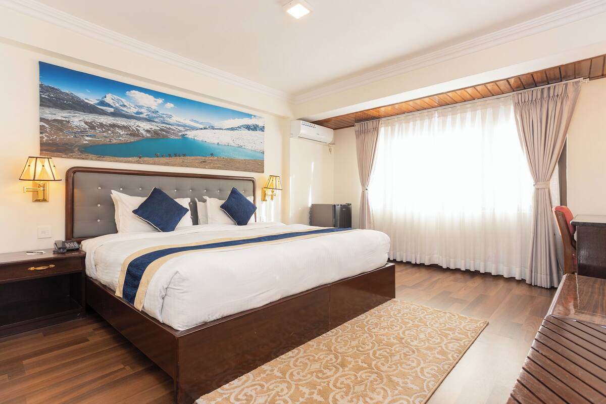 Une chambre d’hôtel avec un grand lit, une table de chevet, un bureau, une chaise, une grande fenêtre avec des rideaux et un tableau représentant un paysage de montagne.
