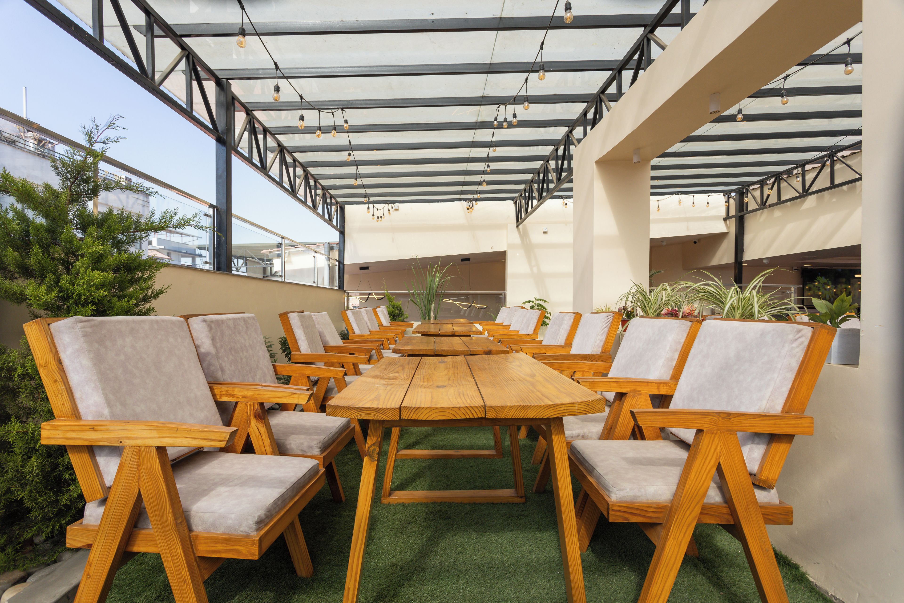Un espacio al aire libre con mesas y sillas de madera, techo de vidrio y césped artificial verde.