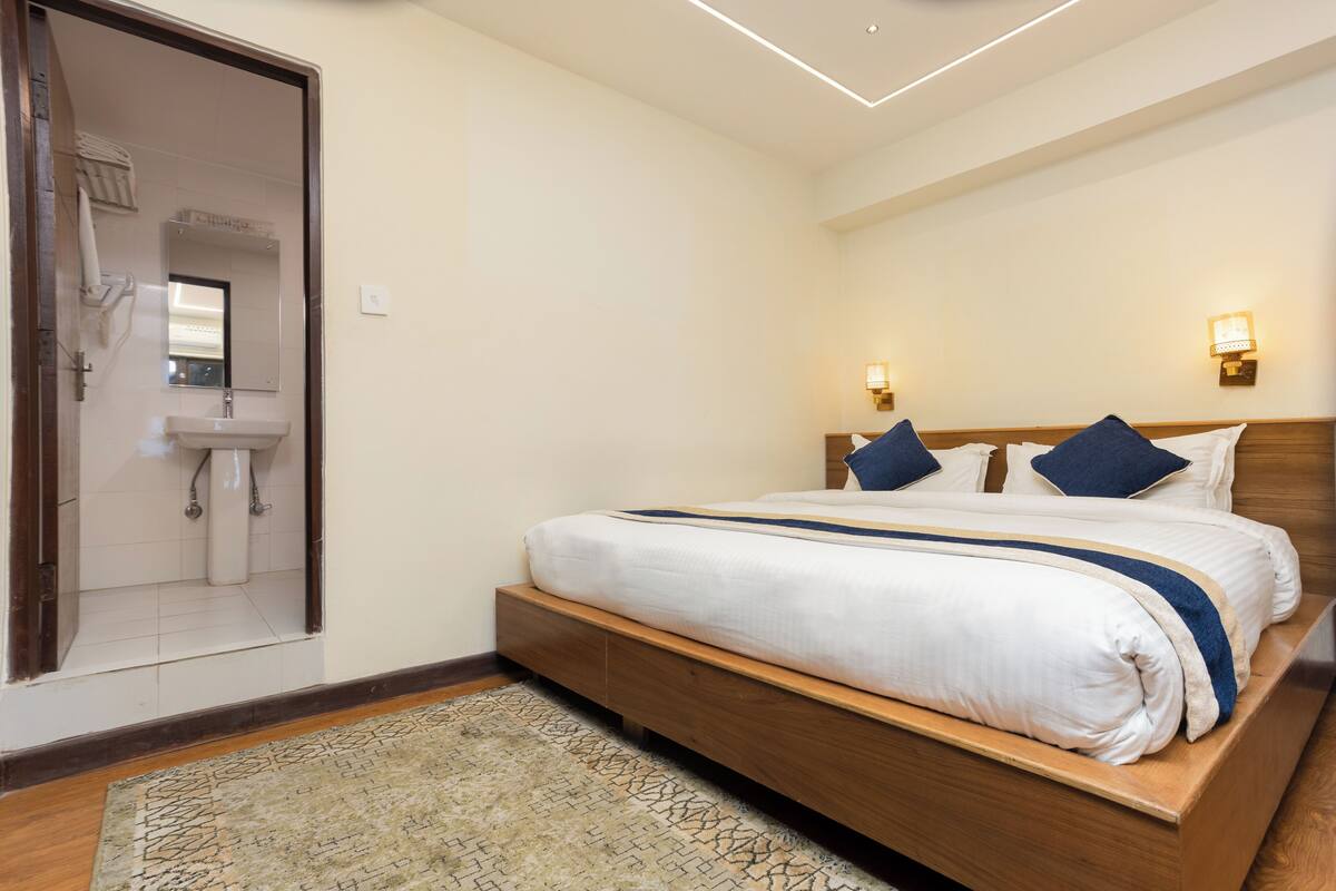 Une chambre avec un lit en bois, du linge de lit blanc, un tapis à motifs, une salle de bain avec un lavabo et un miroir, et des lampes fixées au mur.