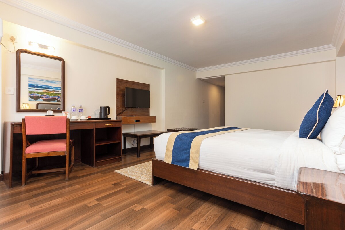 Habitación de hotel con escritorio de madera, una silla, cama con cubrecama azul y beige, televisor de pantalla plana montado en la pared y una mesita de noche con lámpara.