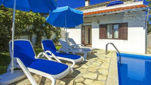 Un espace aménagé au bord de la piscine, avec des chaises longues bleues et des parasols, une terrasse en pierre et un bâtiment blanc aux volets bruns.