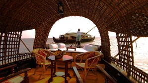 Interior de barco com teto de palha, móveis de vime e vista para outro barco na água.