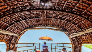 Interior de barco com mesa central, duas poltronas reclináveis, uma mesinha com bebida e uma pessoa sob um guarda-sol.
